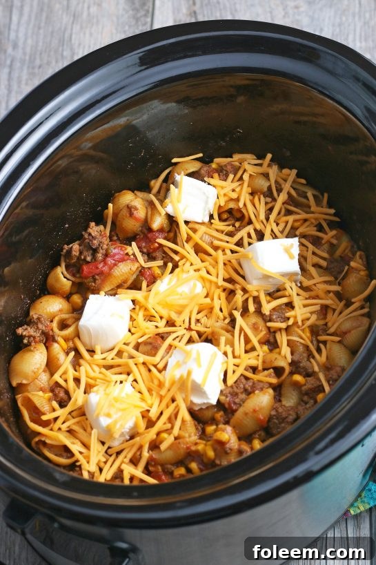 Shredded cheddar cheese being sprinkled generously over the top of the finished slow cooker Mexican pasta, adding a final touch of cheesy deliciousness.