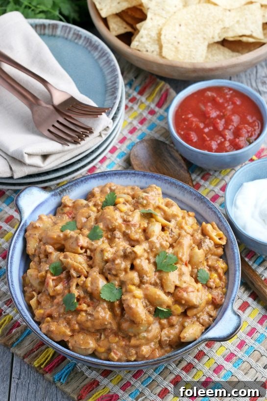 A beautifully set dinner table with a bowl full of Mexican pasta salad, accompanied by a basket of tortilla chips, salsa, and silverware, inviting diners to enjoy.