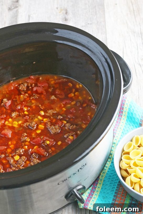 The final step before cooking: adding medium pasta shells to the slow cooker for the Mexican pasta salad, ensuring they are mixed in with the other ingredients.