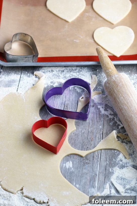 Heart-shaped cookie cutters are actively being used to cut out dough rolled to a perfect thickness, preparing the cookies for baking.