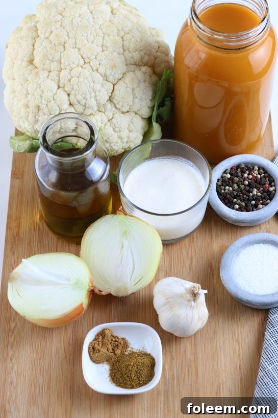 A vibrant flat lay of all the fresh ingredients needed to make homemade roasted cauliflower soup, including a head of cauliflower, onions, garlic, spices, vegetable broth, and heavy cream.