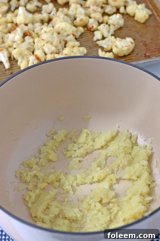 A close-up shot of diced yellow onions and minced garlic sizzling in olive oil in a pan, releasing their aromatic flavors, a crucial step before adding spices for the cauliflower soup.