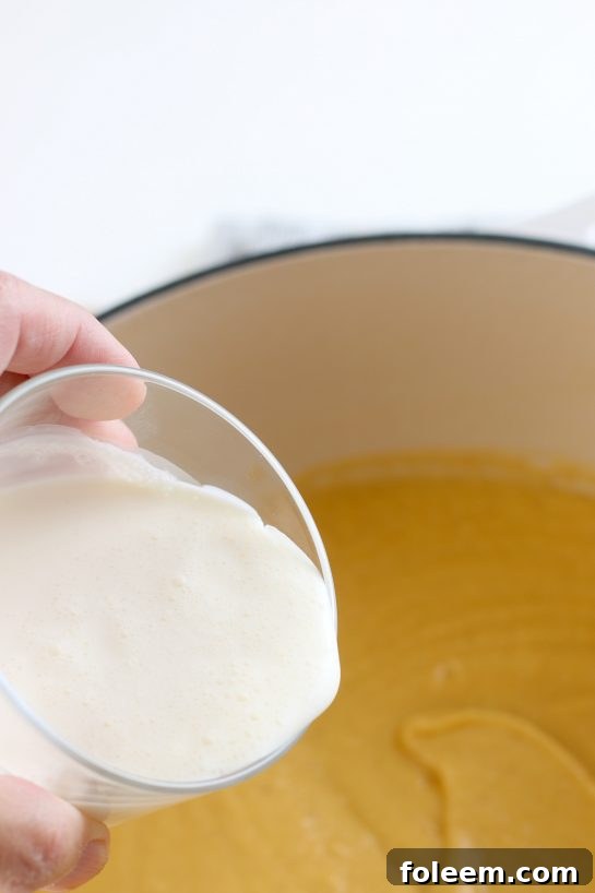 A stream of heavy cream being poured into the blended roasted cauliflower soup in a pot, creating a swirl of white and yellow, signifying the final step of achieving ultimate creaminess.