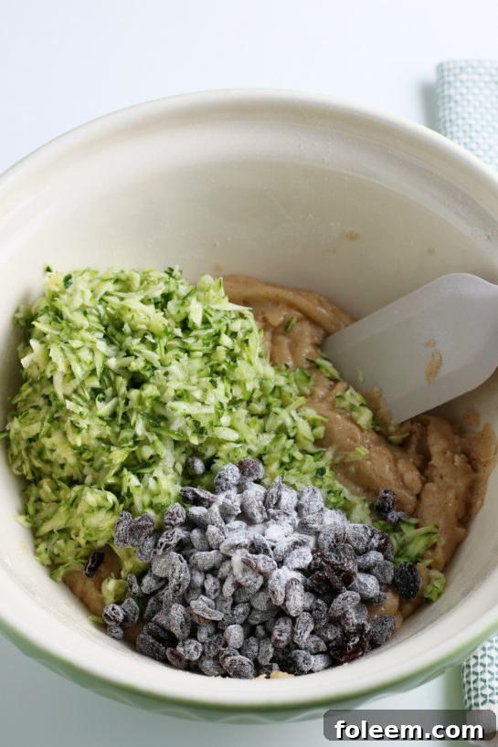 Shredded zucchini and optional raisins being mixed gently into the muffin batter, ensuring even distribution of these key ingredients for ultimate moisture and flavor.
