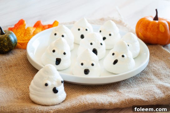 A festive display of cute Halloween Boo Meringues, decorated with candy eyes and black icing for mouths, on a serving plate.