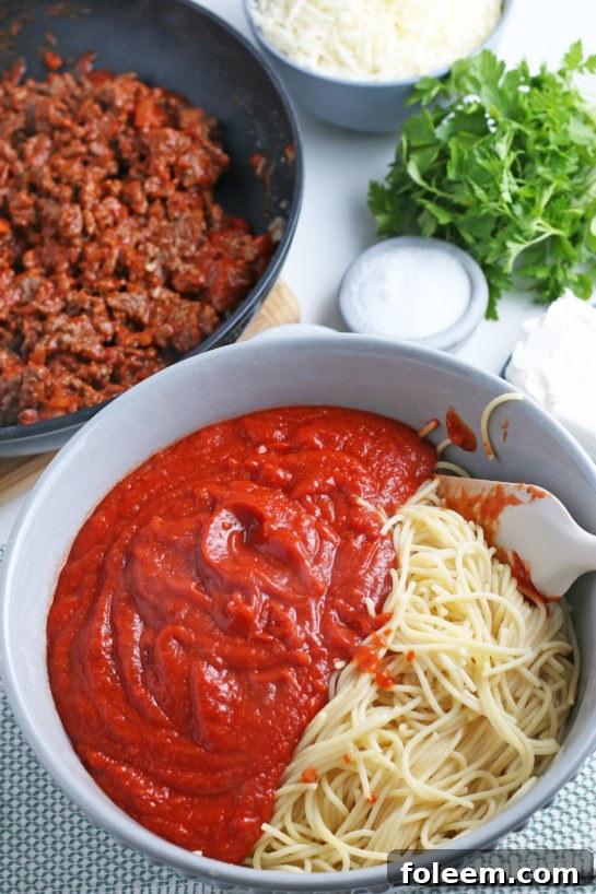 Al dente spaghetti is gently mixed into the aromatic meat sauce, ensuring every strand is coated before layering.