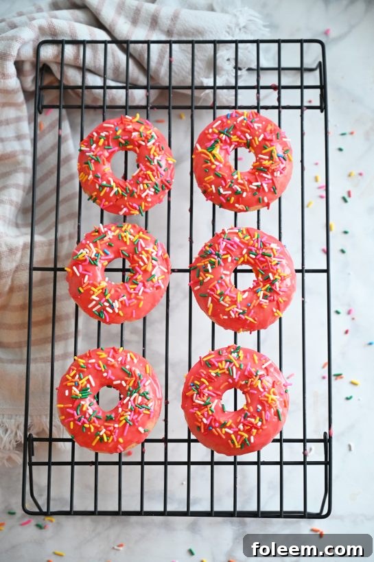 Overhead shot of the finished Vanilla-Glazed Baked Donuts recipe with vibrant pink glaze and colorful sprinkles, arranged neatly on a cooling rack.