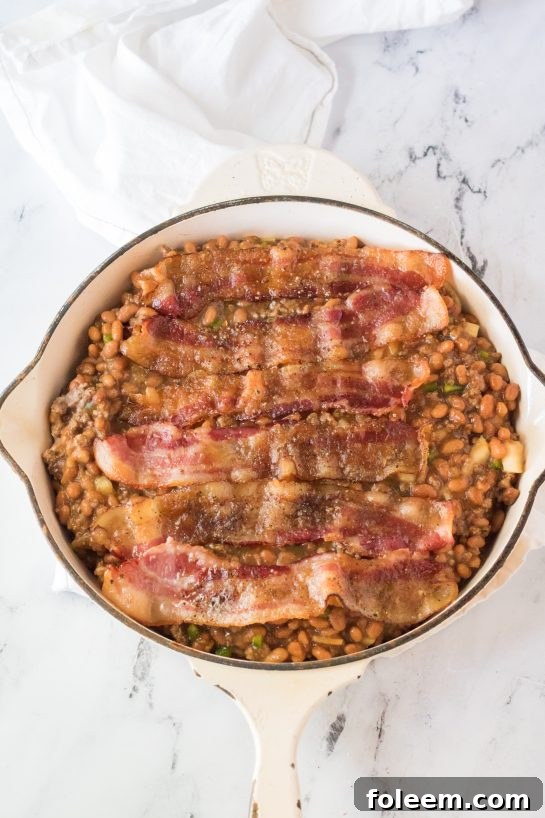 Cooked, crispy peppered brown sugar bacon being placed carefully on top of the simmering Beefy Baked Beans in an oven-safe skillet, ready for final baking.
