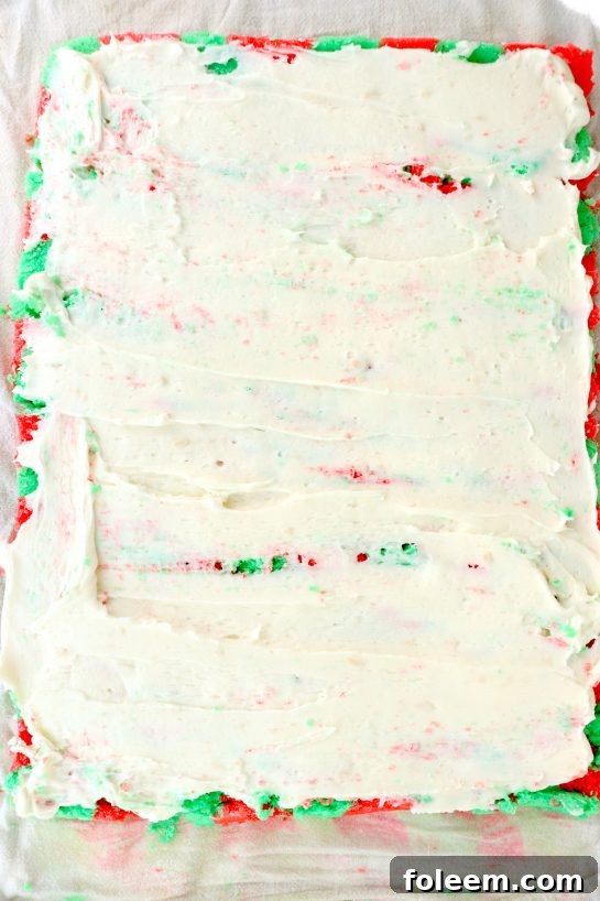 Spreading the creamy vanilla frosting evenly over the baked Christmas cake, preparing it for the rolling process to create a delicious spiral dessert.