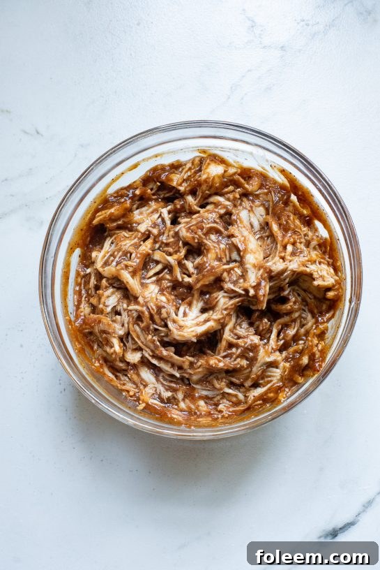 Shredded chicken being mixed thoroughly with rich, sweet, and smoky BBQ sauce in a bowl, a key step for the Sheet Pan BBQ Chicken Pizza.