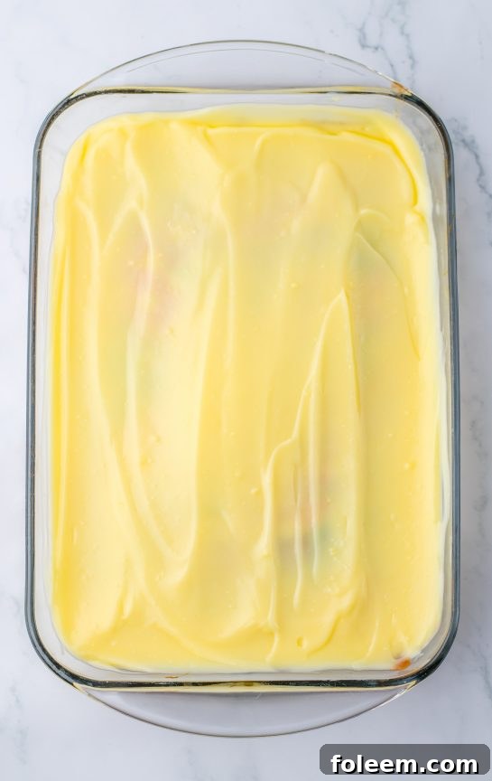 Spreading a thick layer of white chocolate pudding over the pastel-colored, hole-poked Easter Poke Cake.
