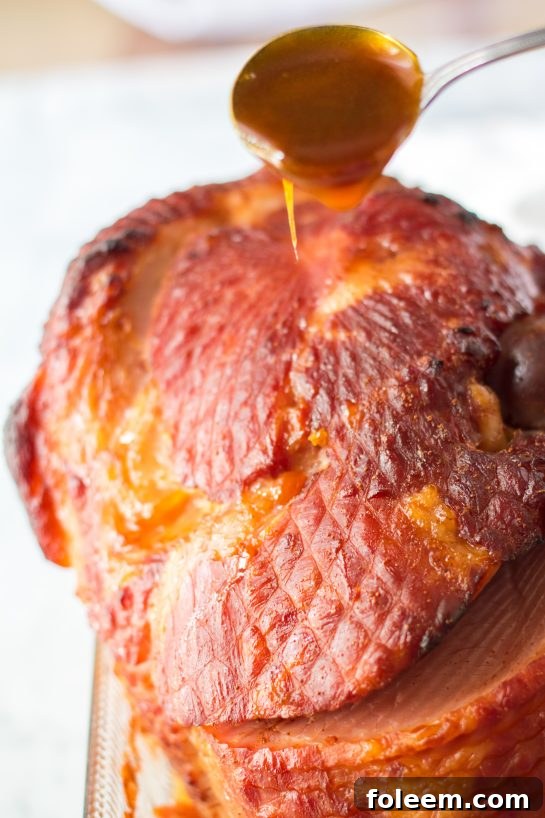 Thick orange, brown sugar, and honey glaze being poured generously over a spiral-sliced Christmas ham in a roasting pan.