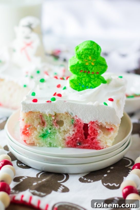 A close-up of a Christmas Poke Cake slice, showing the vibrant red and green Jell-O streaks within the white cake, topped with whipped cream and sprinkles.