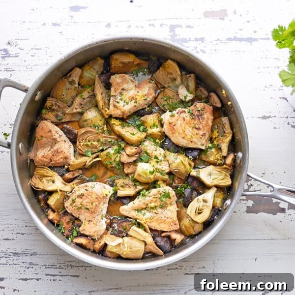 Skillet of Braised Chicken, Mushrooms and Baby Artichokes set on a white, wooden table.