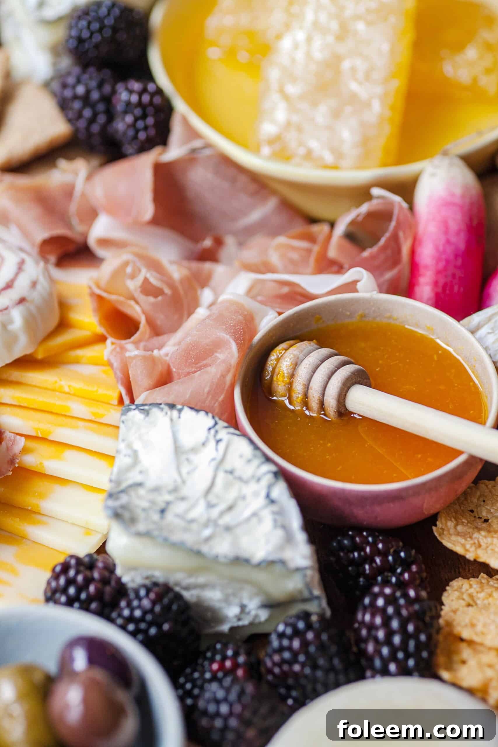 Bowl of honey next to meat, cheese, fruit, and other grazing board foods.