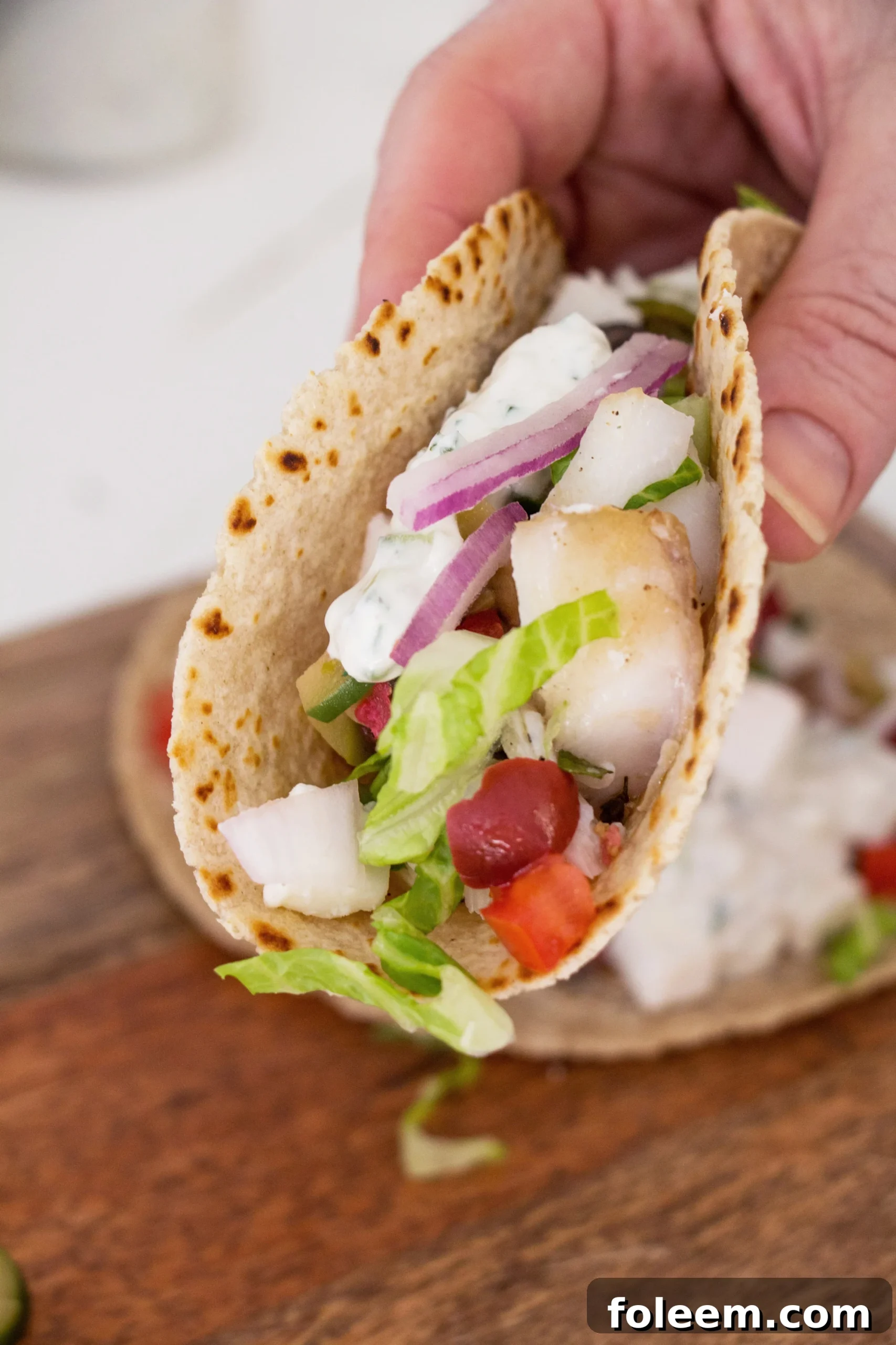 Person holding a Greek Fish Taco stuffed with ingredients such as feta and red onions.