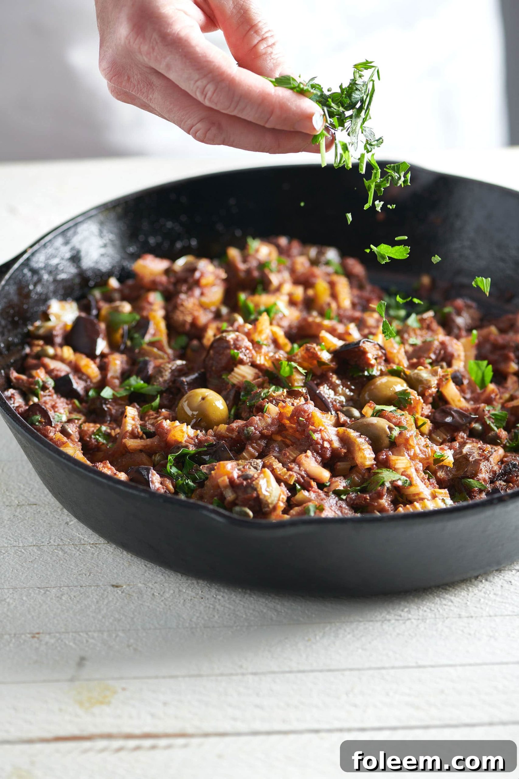 Caponata in a cast iron skillet being sprinkled with fresh parsley.