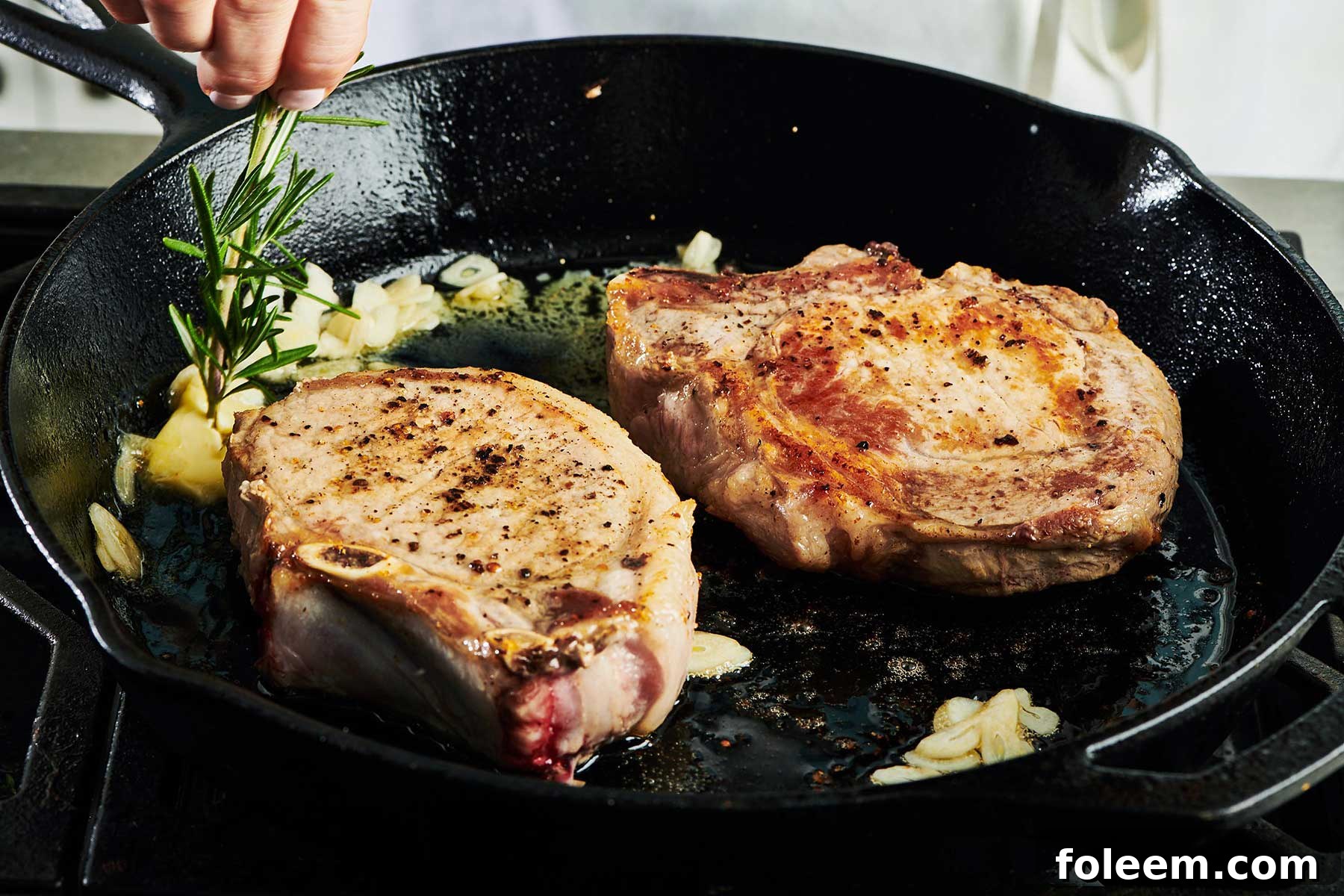 Adding fresh rosemary to pork chops in cast iron pan on stove.