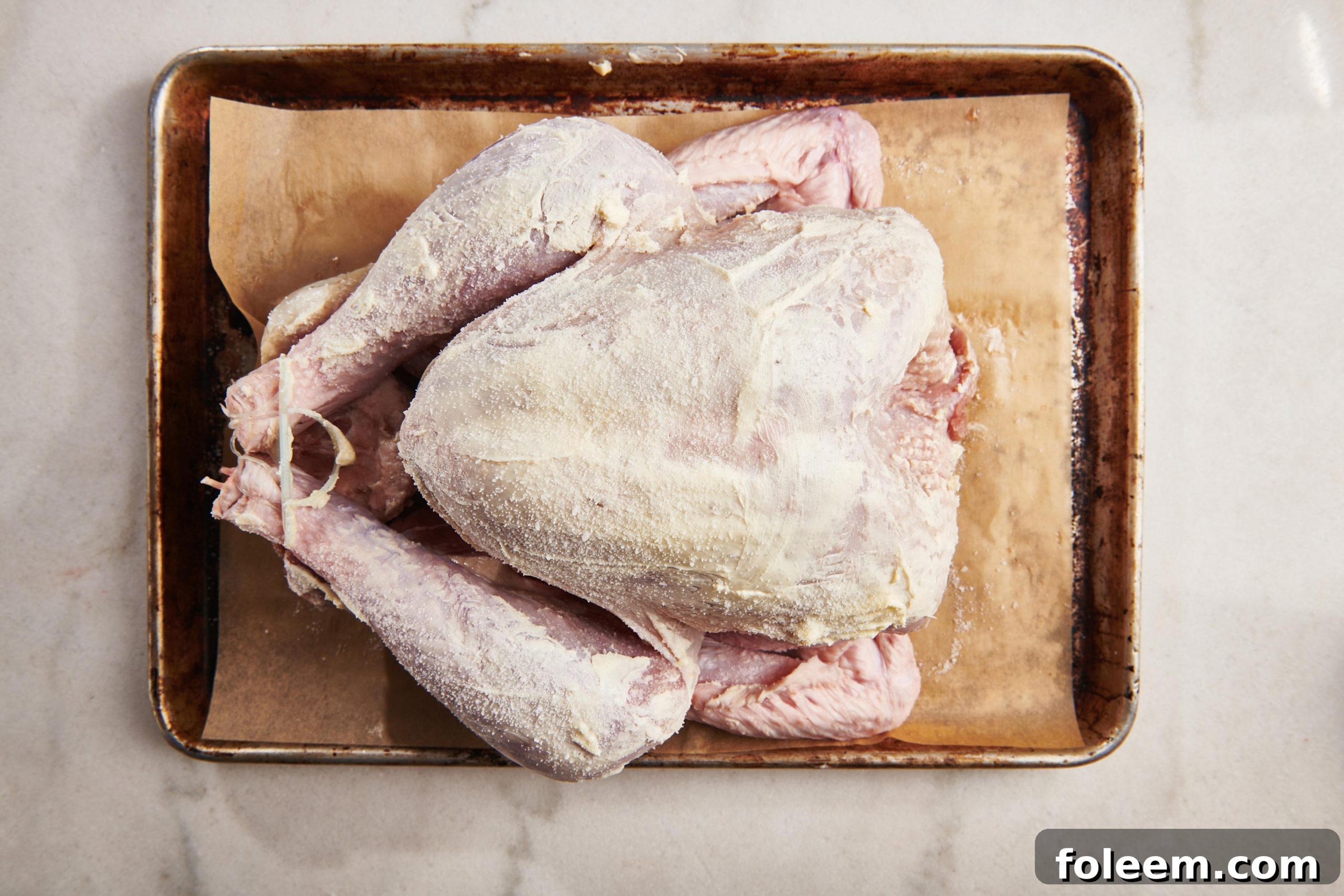 Uncooked trussed turkey smeared with butter on a baking sheet.