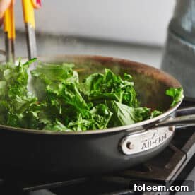 Stirring broccoli rabe with tongs as it cooks in large pan on stove.