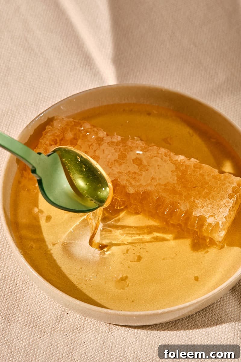 Green spoon dipping into bowl of honey with a fresh honeycomb.